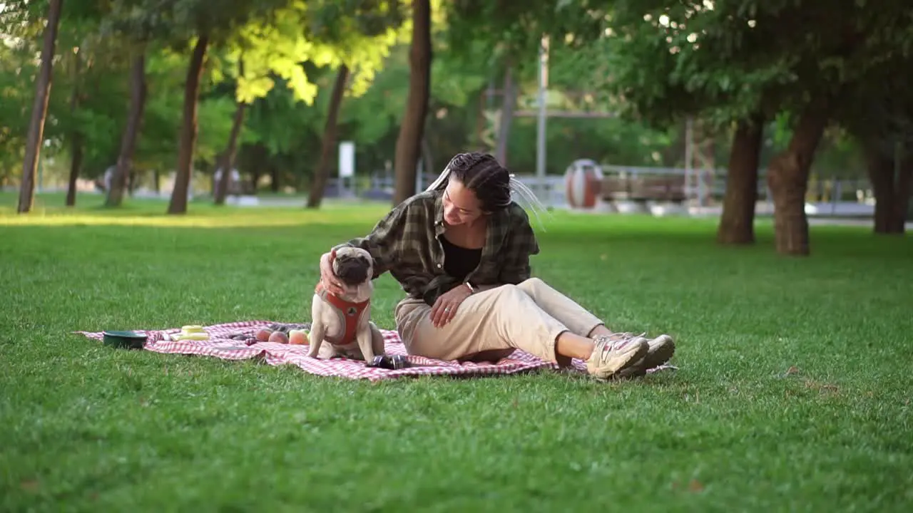 Girl Sitting On Plaid On Lawn In A Park While Dog Sitting In Front Of Her She Pets Him