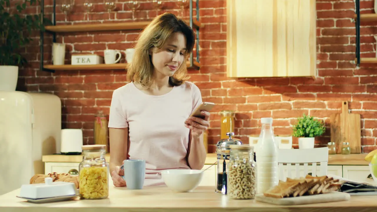 Pretty Young Woman Having Breakfast In The Morning In The Kitchen And Chatting Scrolling And Tapping On The Smartphone