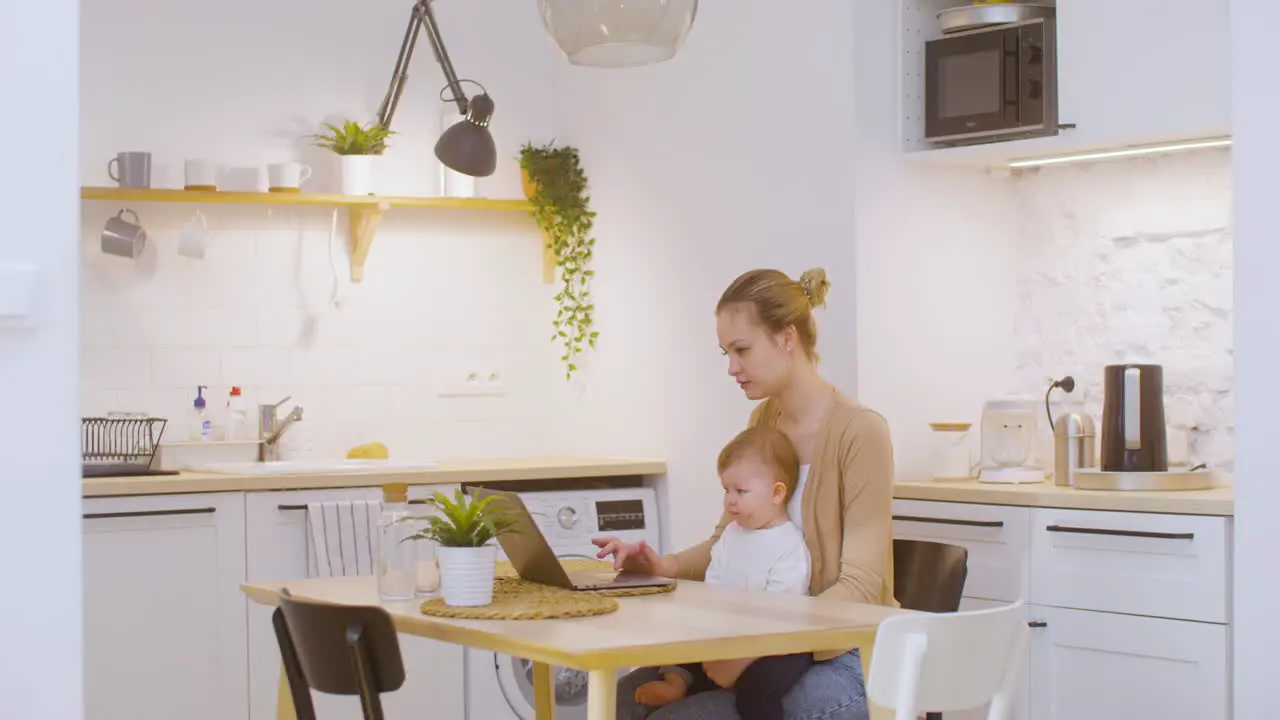 Young Woman Working On Laptop Computer While Sitting With Baby Boy At Home