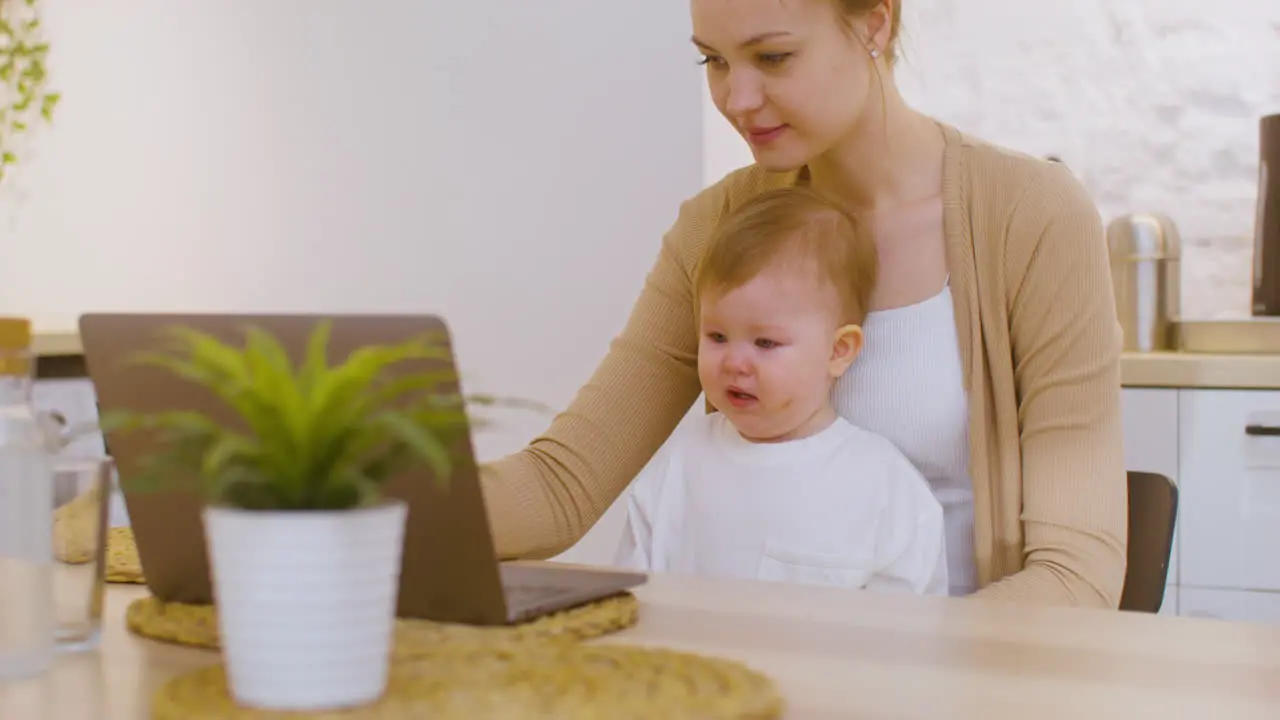 Young Woman Working On Laptop Computer While Sitting With Baby Boy At Home 1