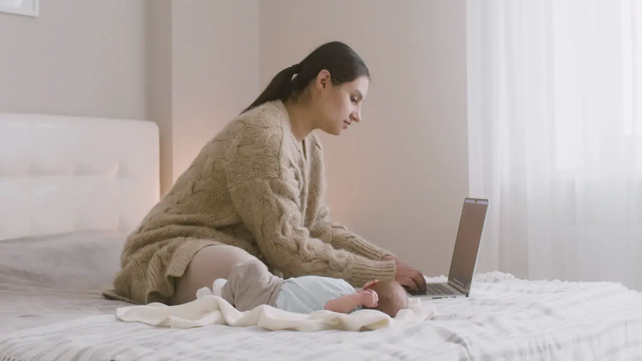 Young Mother Sitting On The Bed And Working On Laptop Computer While Her Newborn Baby Lying Next To Her