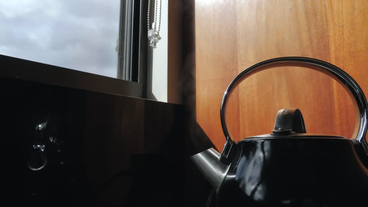 Wide shot of black household domestic kitchen kettle slowly boils for cup of tea coffee with steam water vapor coming out of metal spout with window in background