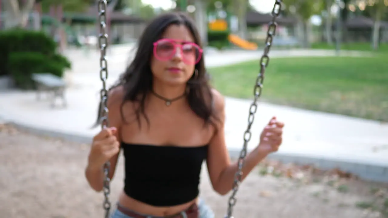 Close up on a pretty hispanic girl playing on the playground swing set wearing retro pink sunglasses in slow motion