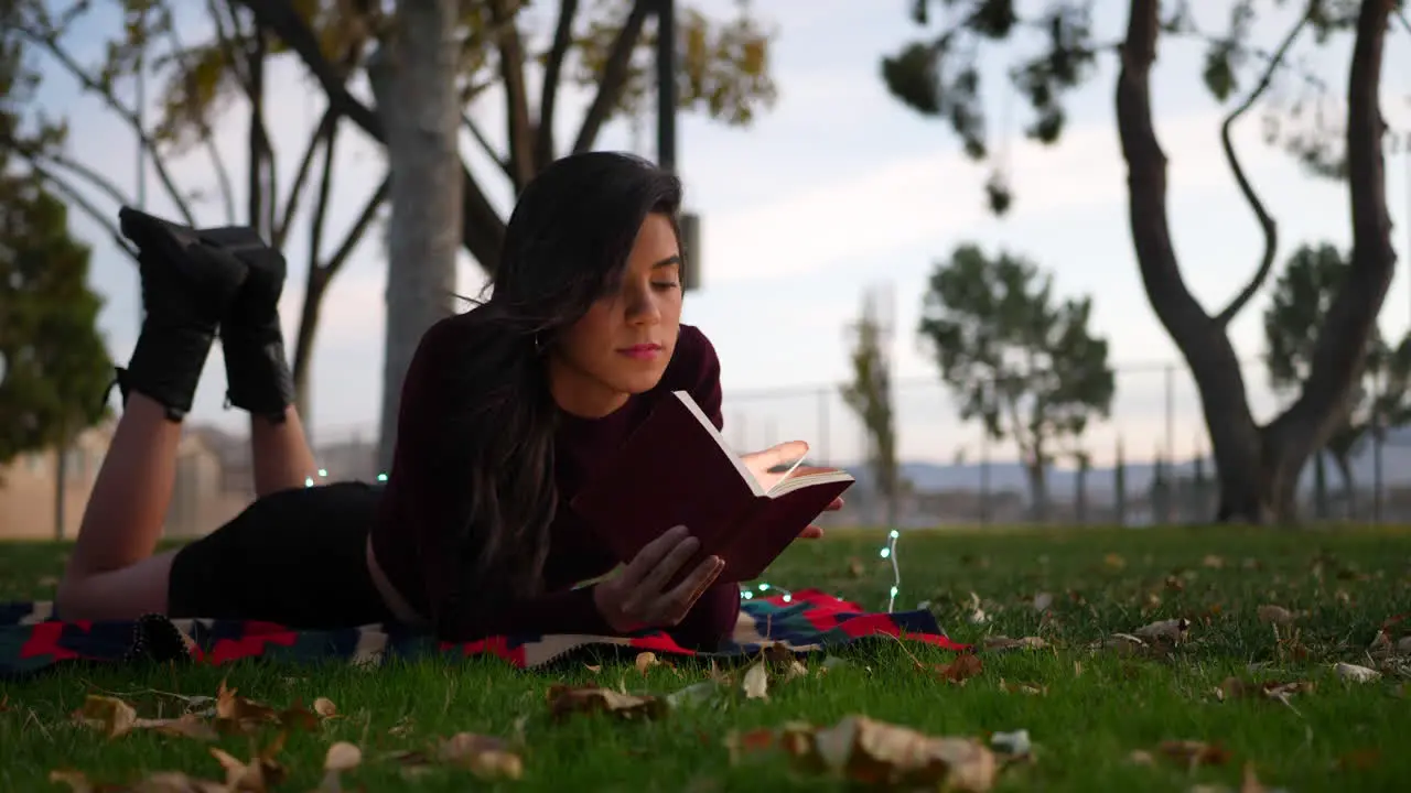 A young adult woman reading a novel or story book in the park at sunset laying in the grass with autumn leaves SLOW MOTION