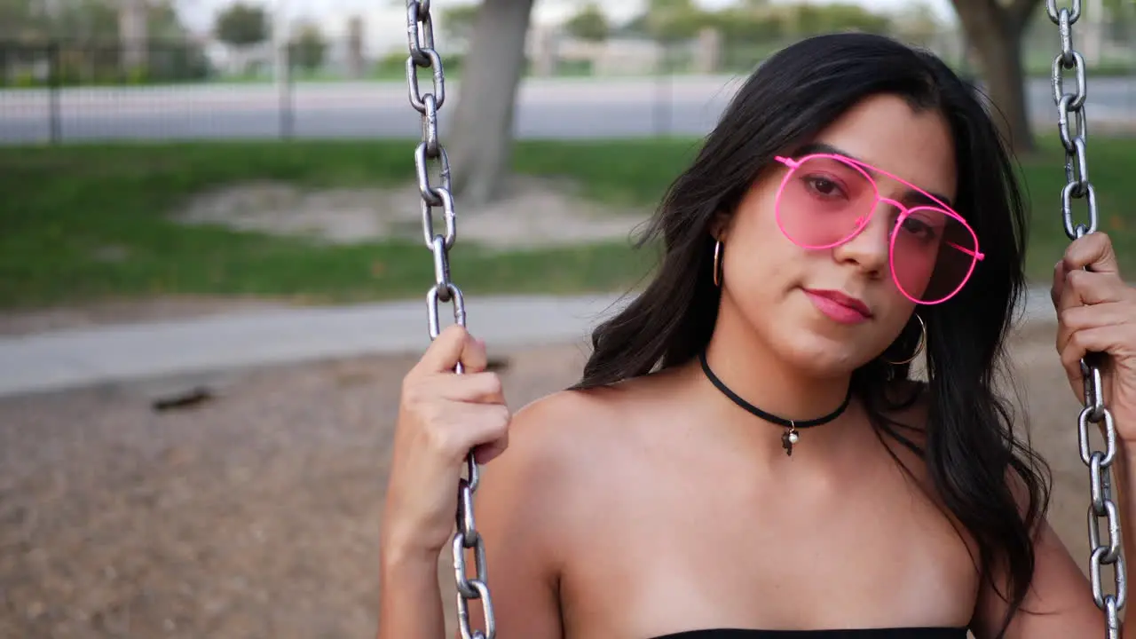 A beautiful young hispanic woman playing and swinging on a park playground swing set wearing retro pink sunglasses in slow motion