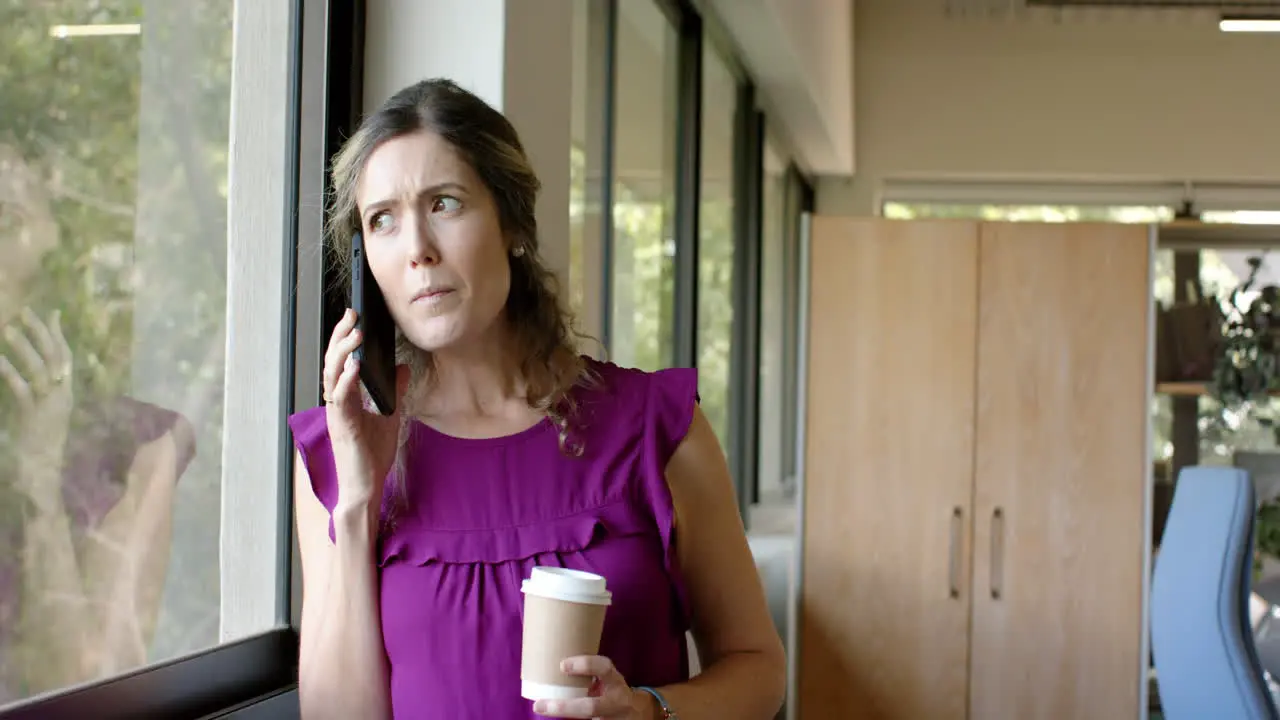 Caucasian casual businesswoman drinking coffee and talking on smartphone in office slow motion