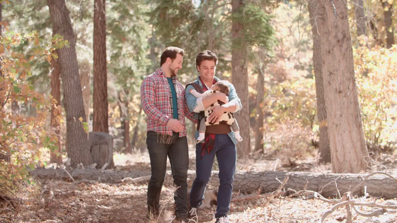 Male couple in forest with baby in sling walk towards camera