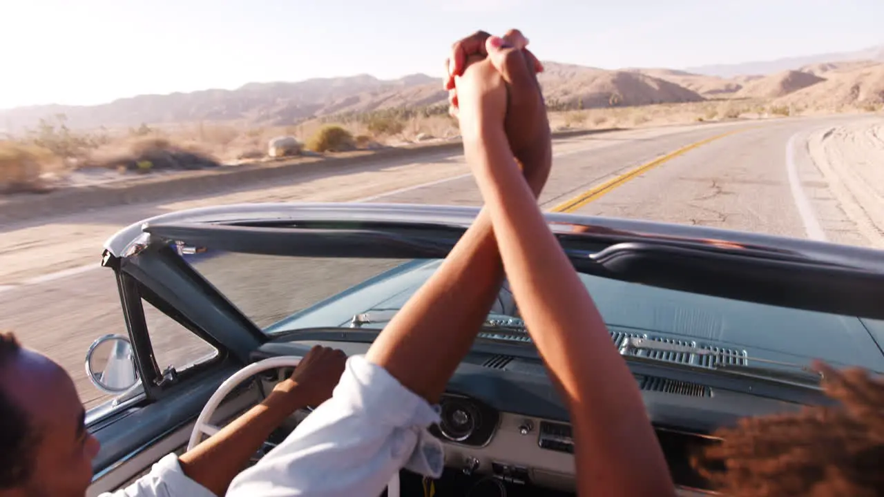 Couple in open top car hold hands in the air elevated view