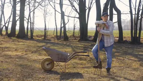 Caucasian man leaning on the stick of a rake standing near a wheelbarrow in the countryside