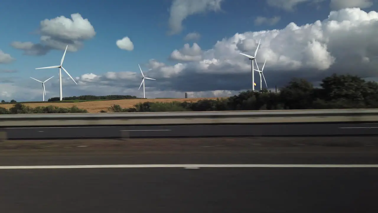 Wind farm from seen from a road with burning fossil fuel gas flaring from a chimney in the distance along side green fields