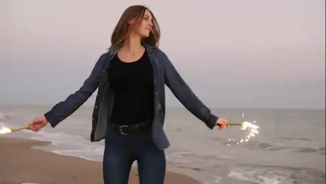 Young beautiful woman running by the sea during sunset and holding burning sparkling candles in both hands turning around