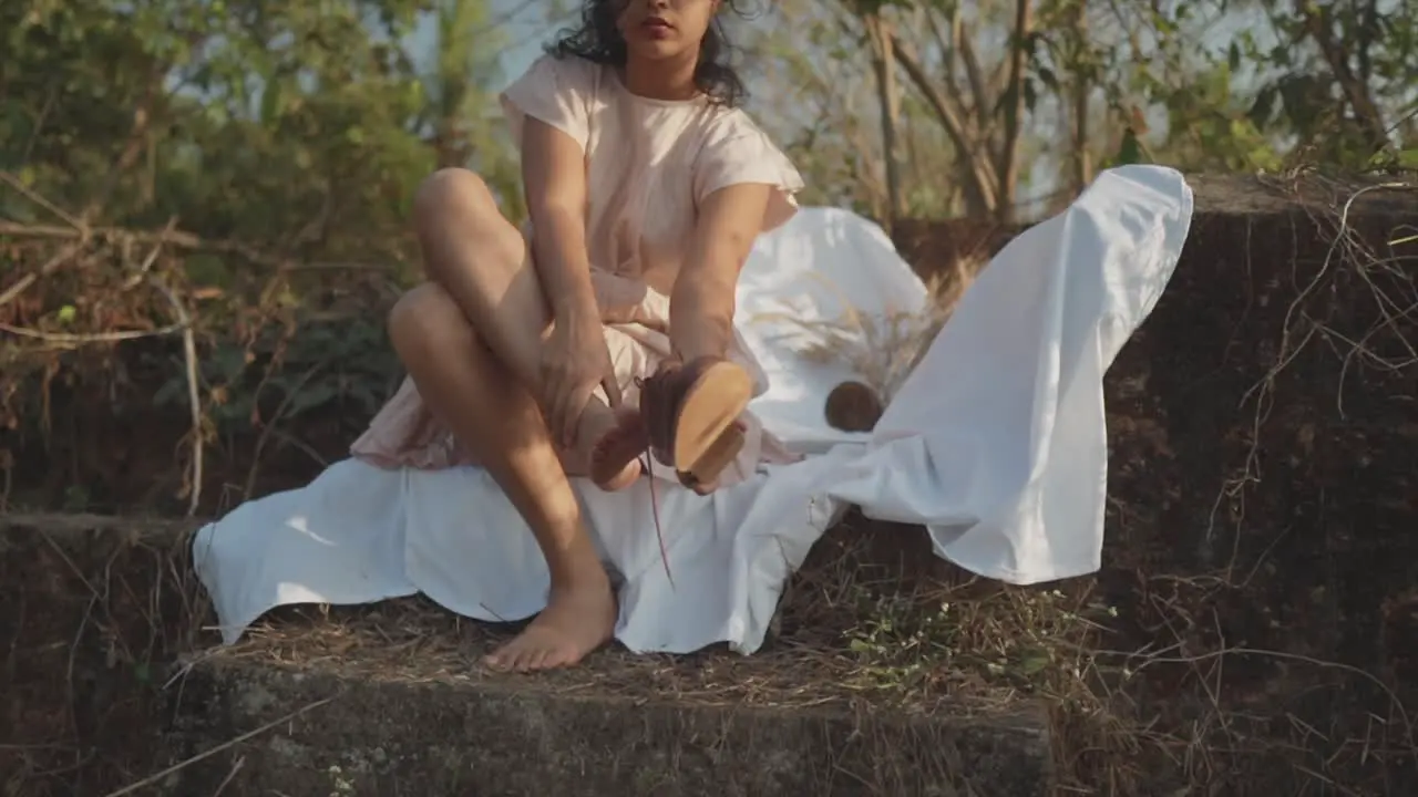 Slow motion handheld panning shot of a beautiful indian model sitting on a white sheet in the garden taking off her shoe on a sunny day