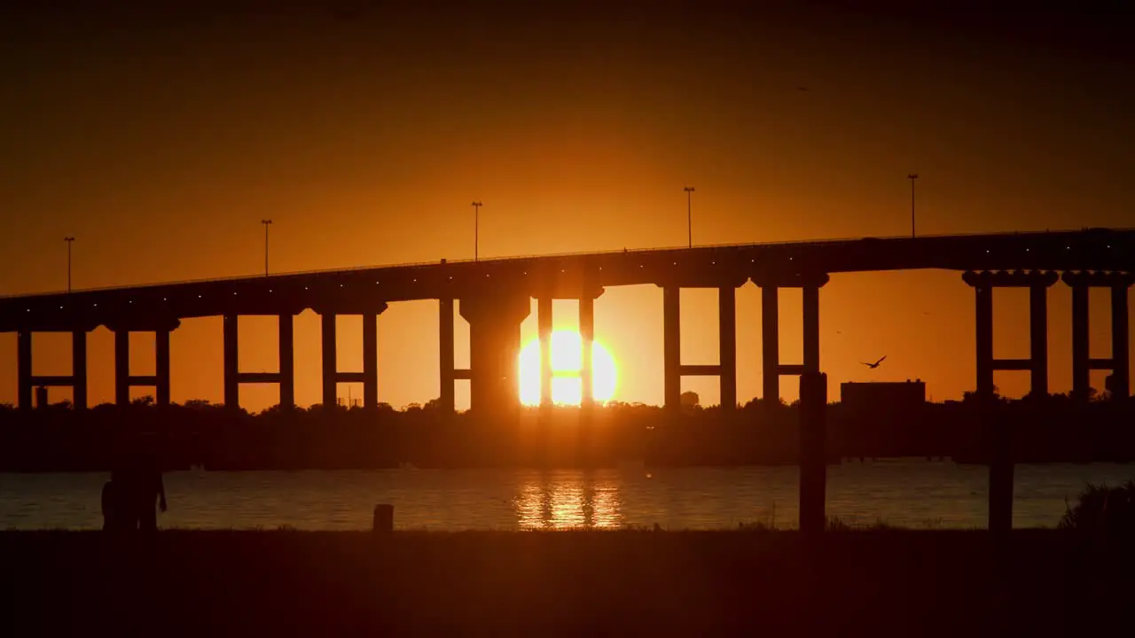 A slow motions sunset under a bridge over water while people walk through the foreground