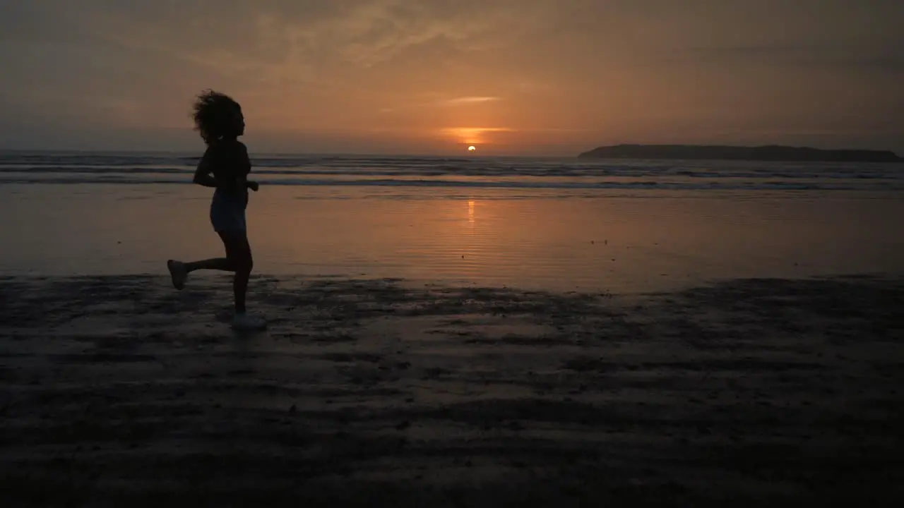 Landscape view of a silhouette of a young woman running on a sandy beach by the ocean at sunrise