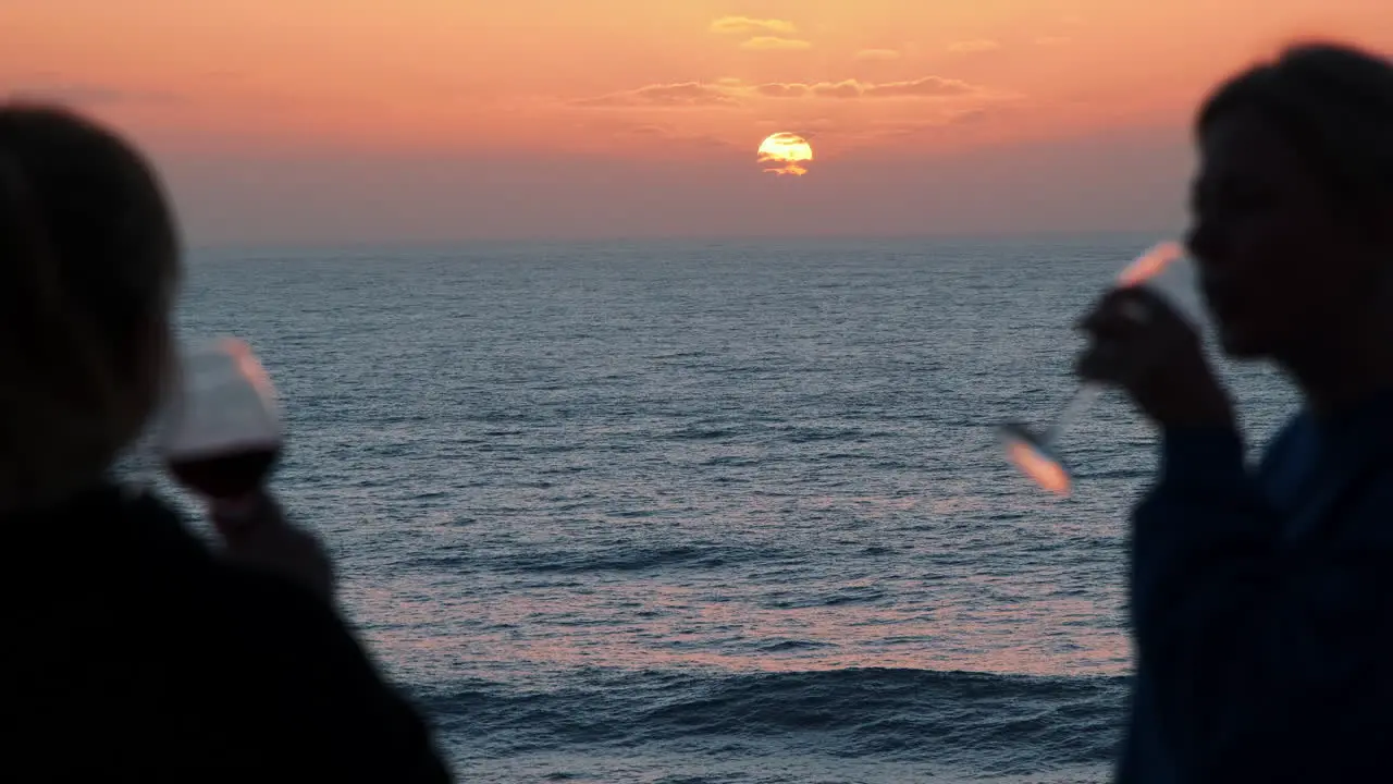 Silhouette of two girls drinking a red wine in front of beautiful ocean and golden sunset in background