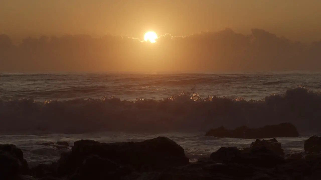 Slow motion shot of orange gold sun rising over low cloud and breaking surf