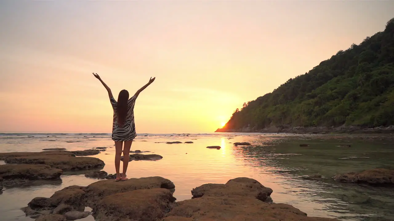 Asian Woman standing on rock by the sea water and raising hands up when the sun going down behind the mountain