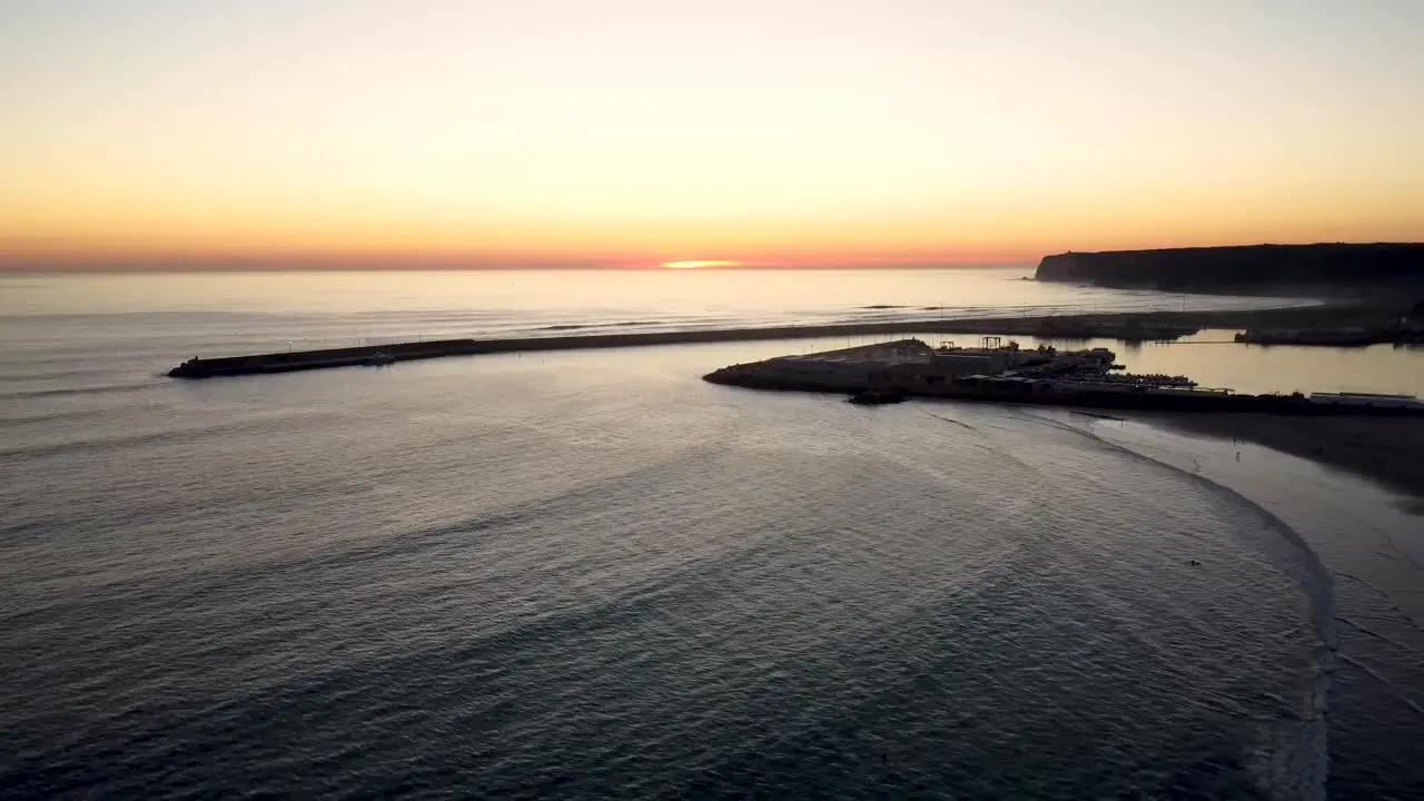 Closing drone shot of waves crashing onto a beach with a fishing port in the background during sunset