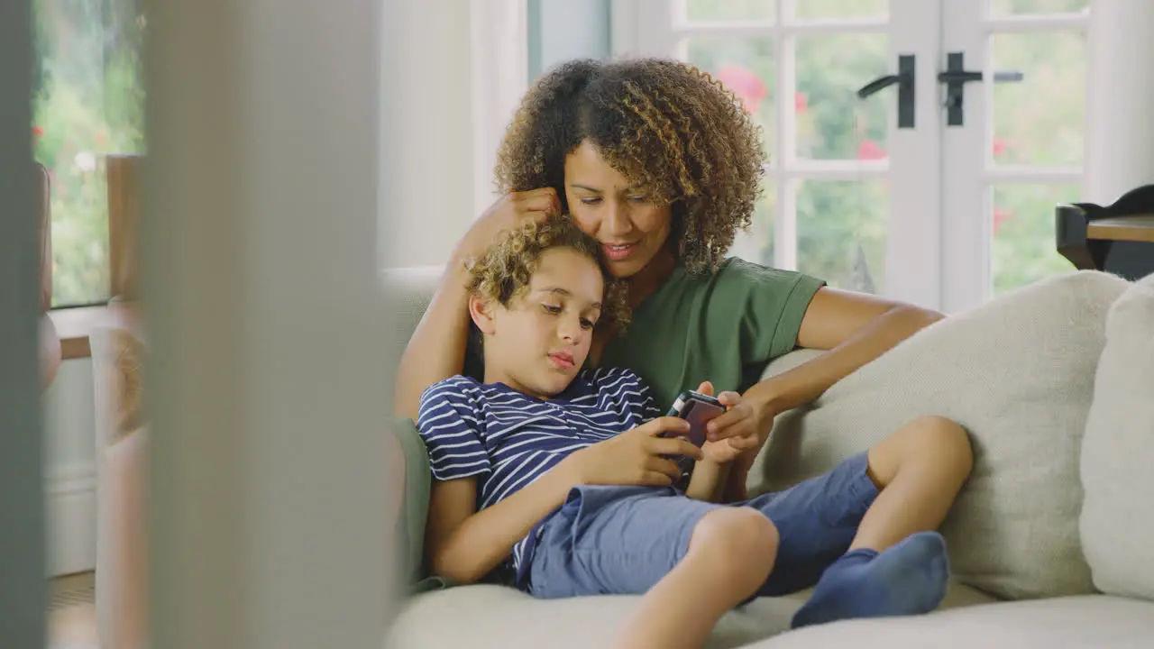 Mother And Son Sitting On Sofa At Home Playing Video Game On Mobile Phone Together