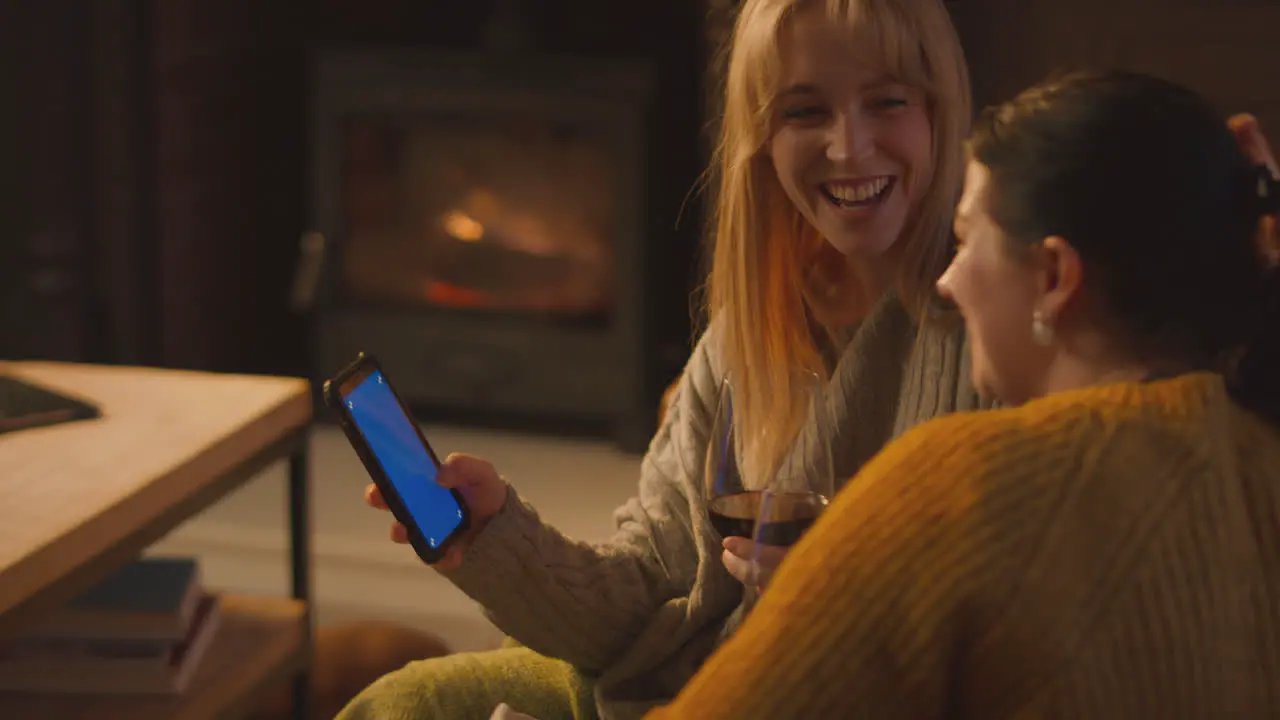 Two Female Friends Relaxing On Sofa Looking At Mobile Phone With Cosy Fire Holding Glass Of Wine