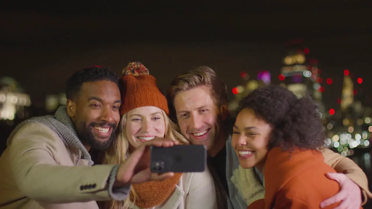 Group of friends wearing winter coats and scarves posing for selfie on mobile phone with city lights in background shot in slow motion