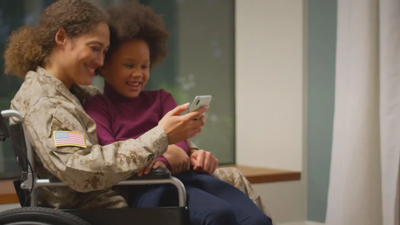 Injured Female American Soldier Wearing Uniform Sitting In Wheelchair Looking At Phone With Daughter