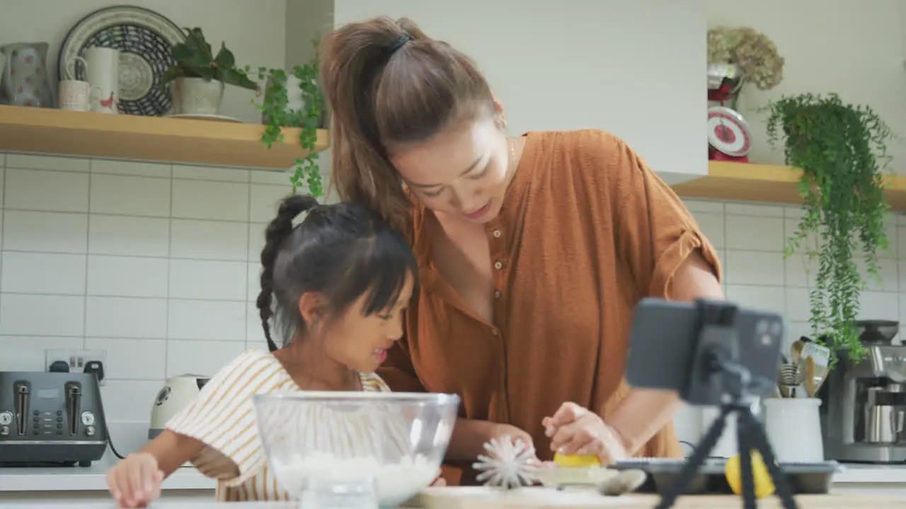 Asian Mother And Daughter Baking Cupcakes In Kitchen At Home Whilst On Vlogging On Mobile Phone