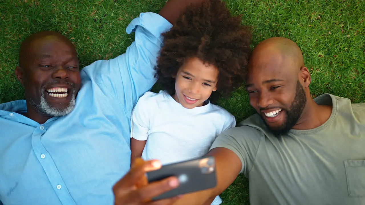 Overhead Shot Of Multi-Generation Male Family Lying On Grass Taking Selfie On Mobile Phone