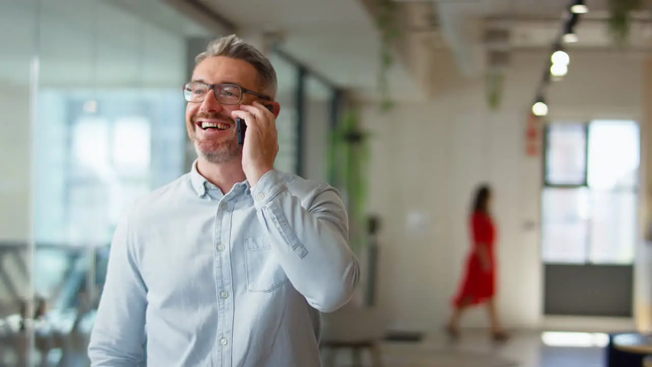 Mature Businessman Standing In Modern Open Plan Office Talking On Mobile Phone