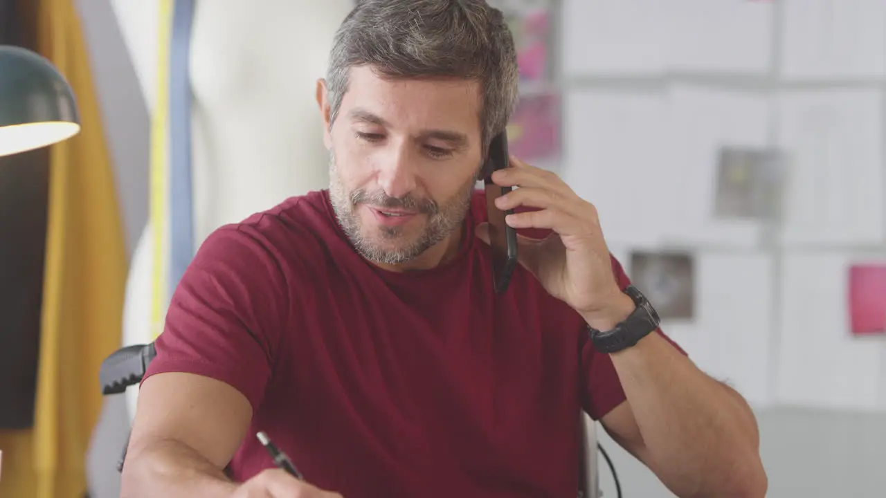 Mature Male Fashion Designer In Wheelchair In Studio Working On Sketches And Talking On Mobile Phone