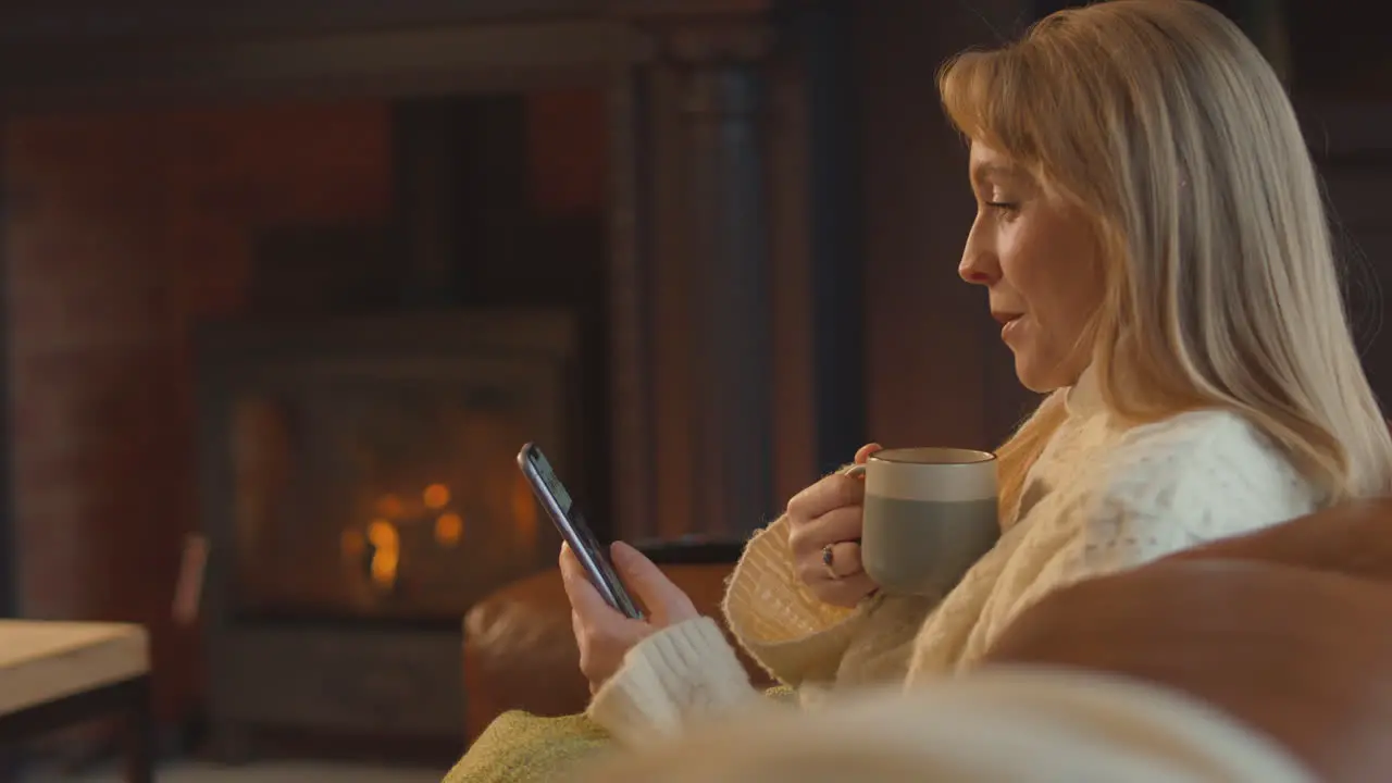 Woman At Home In Lounge With Cosy Fire And Hot Drink Using Mobile Phone