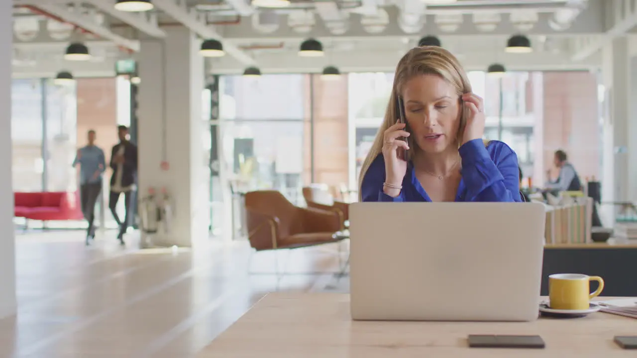 Businesswoman Sitting At Desk On Phone Call In Modern Open Plan Office With Colleagues In Background