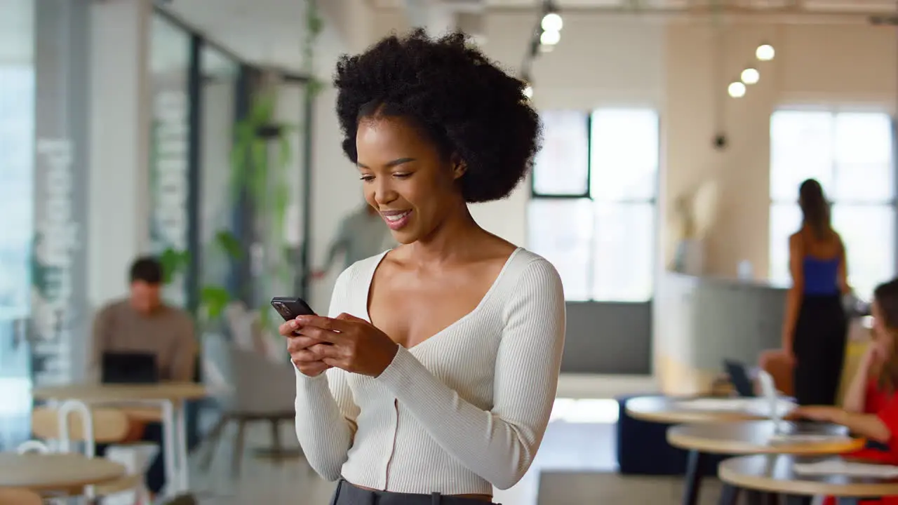 Smiling Businesswoman Standing In Busy Open Plan Office Messaging On Mobile Phone