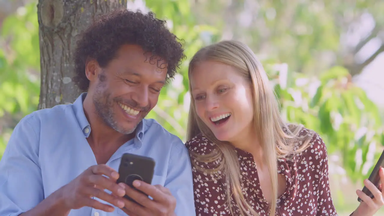 Mature Couple Meeting In Outdoor Park Sitting On Bench Looking At Mobile Phones Together