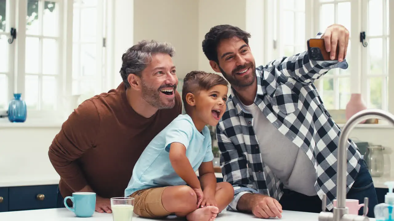 Same Sex Family With Two Dads Taking Selfie In Kitchen With Son Sitting On Counter