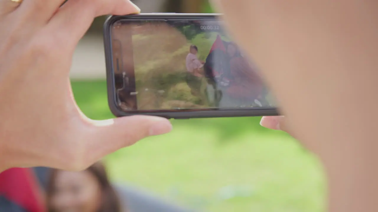 Close Up Of Asian Father Filming Video On Mobile Phone As Family Put Up Tent In Garden At Home