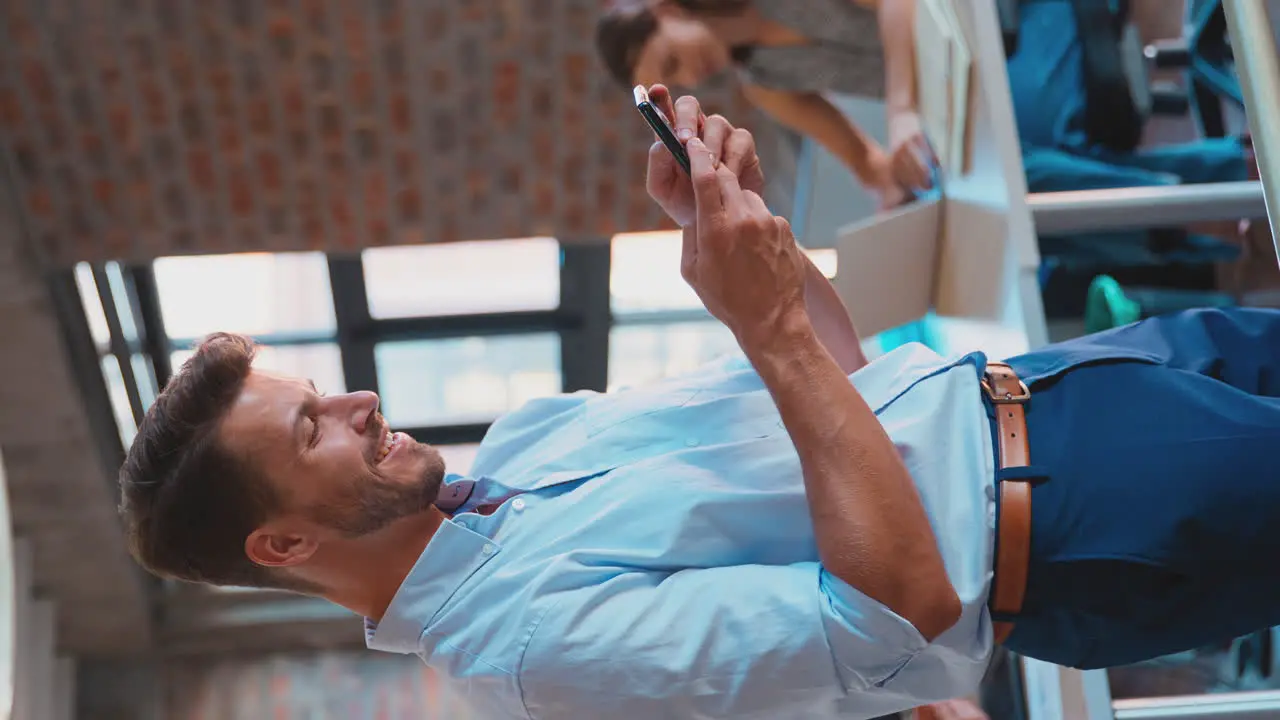 Vertical Video Of Smiling Businessman Standing In Busy Office Messaging On Mobile Phone