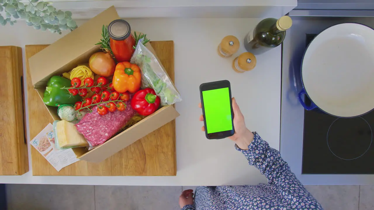 Overhead shot of woman in kitchen with fresh ingredients looking for online recipe on green screen mobile phone shot in slow motion