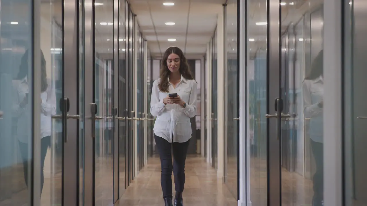 Young Businesswoman Walking Along Corridor Of Modern Office Using Mobile Phone