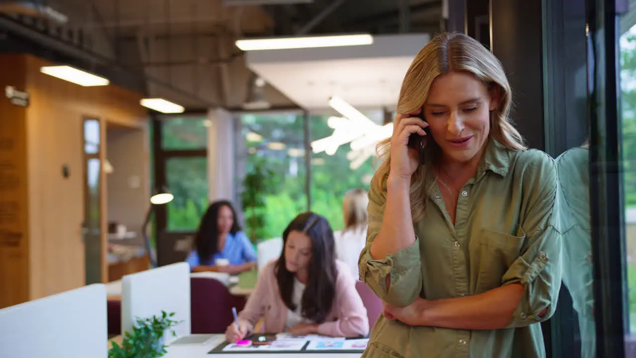 Mature Businesswoman Standing By Window In Busy Office Talking On Mobile Phone