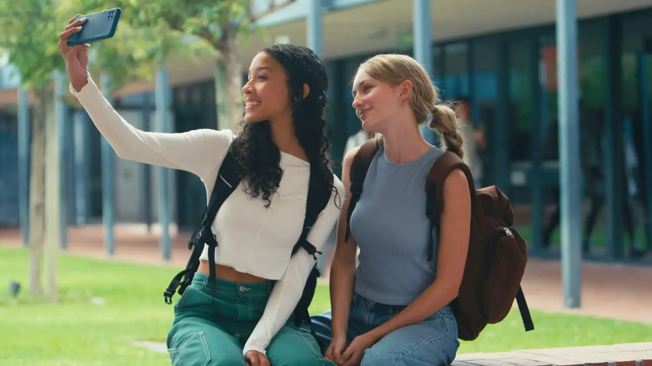 Two Female High School Or Secondary Students Posing For Selfie On Phone Sitting On Wall Outdoors