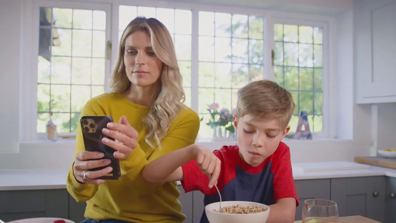 Son At Home Eating Breakfast Cereal At Kitchen Counter As Mother Makes Video Call On Mobile Phone