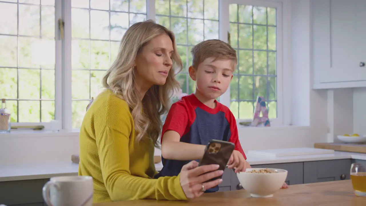 Son At Home Eating Breakfast Cereal At Kitchen Counter Looking At Mobile Phone With Mother