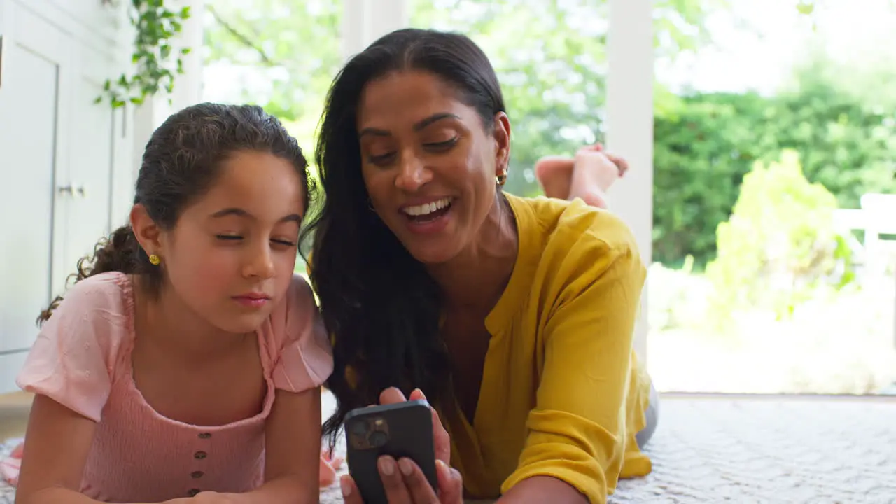 Mother And Daughter At Home Lying On Floor In Lounge Pulling Funny Faces Posing For Selfie On Phone