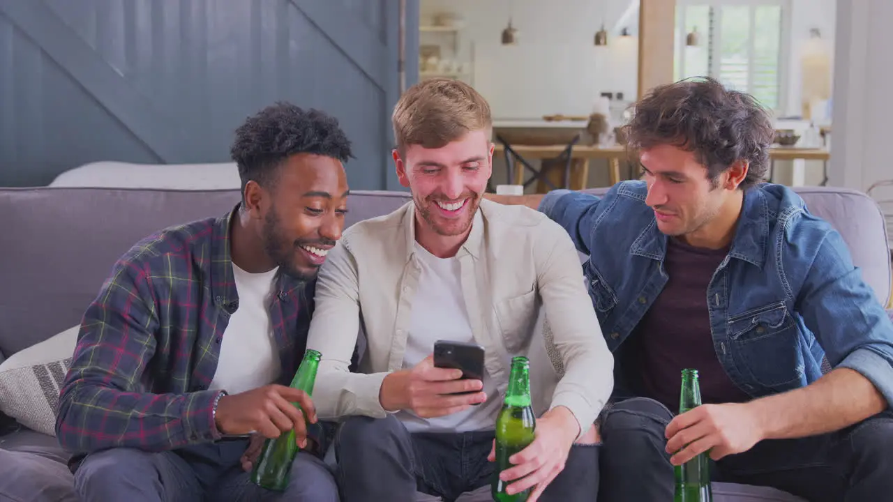 Multi-Cultural Group Of Male Friends On Sofa At Home Drinking Beer And Looking At Mobile Phone