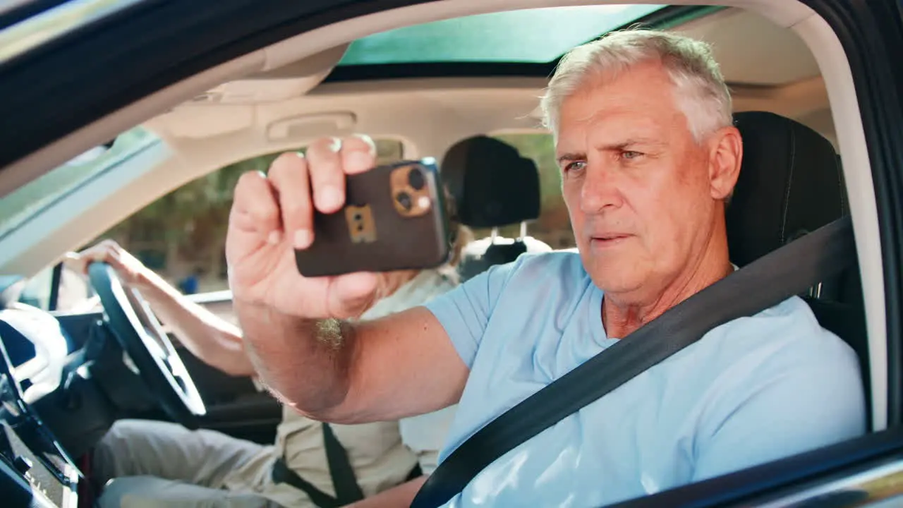 Passenger Taking Photo On Mobile Phone As Senior Couple Enjoy Day Trip Out In Car