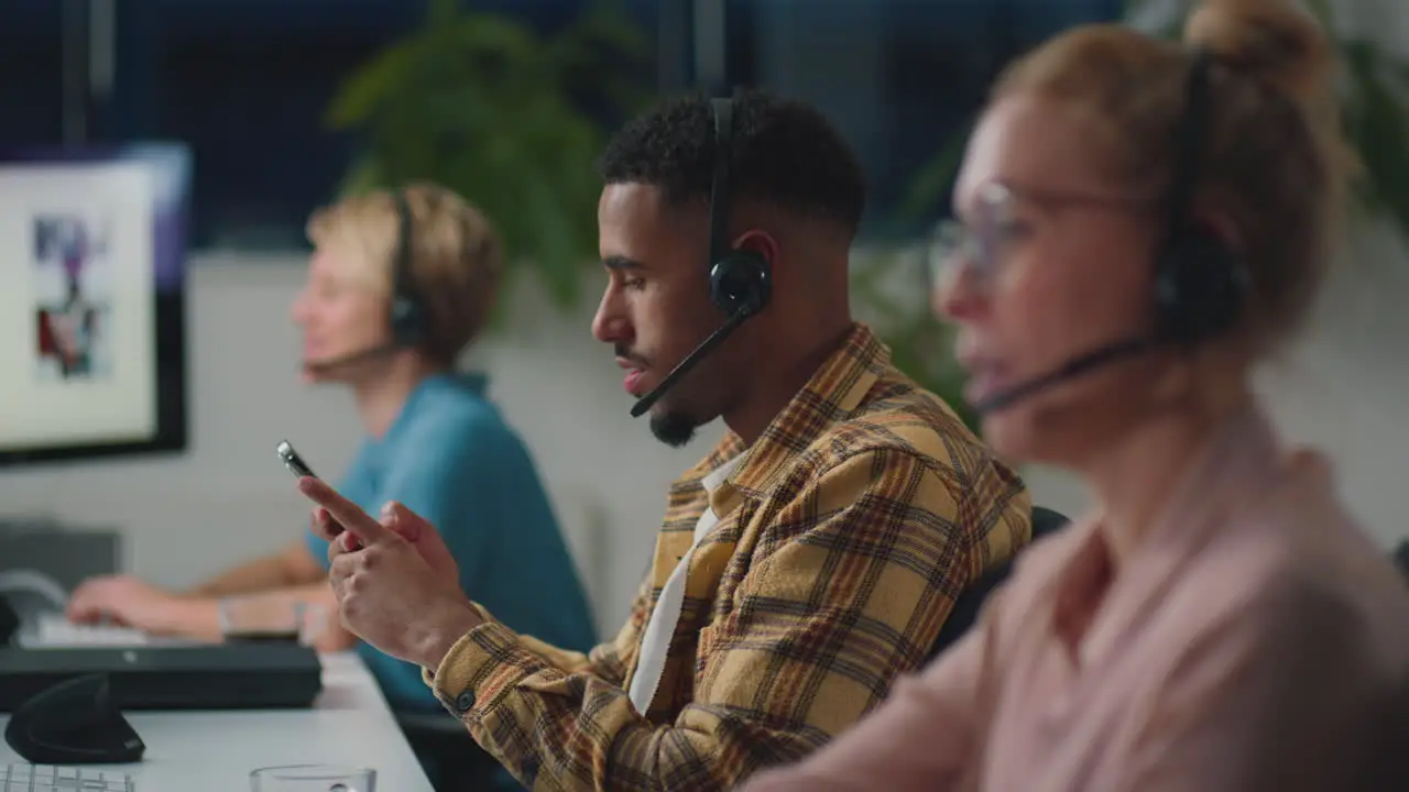 Man In Business Team Wearing Headsets Looking At Mobile Phone Working Late In Sales Or Support