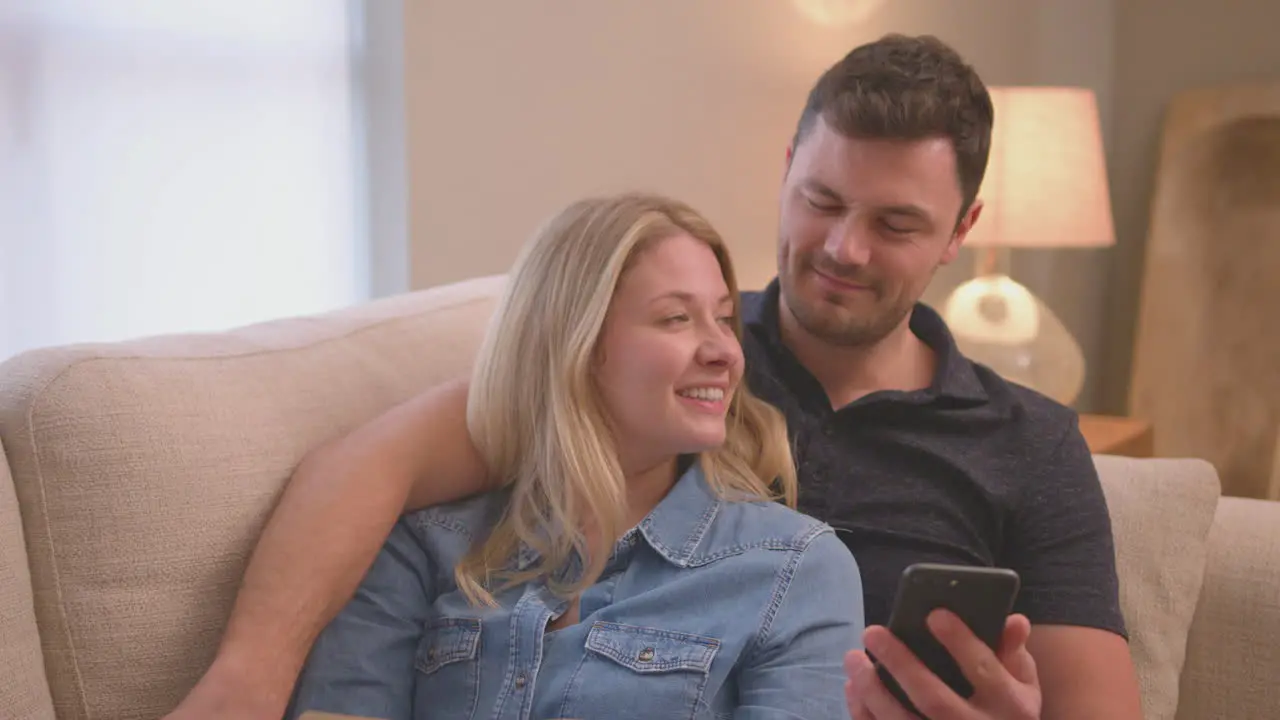 Evening shot of couple relaxing on sofa at home as man looks at mobile phone and woman reads book shot in slow motion