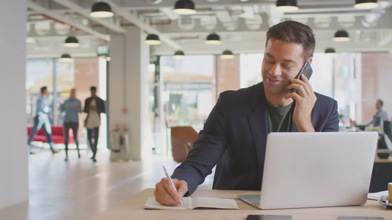 Businessman Sitting At Desk On Phone Call In Modern Open Plan Office With Colleagues In Background