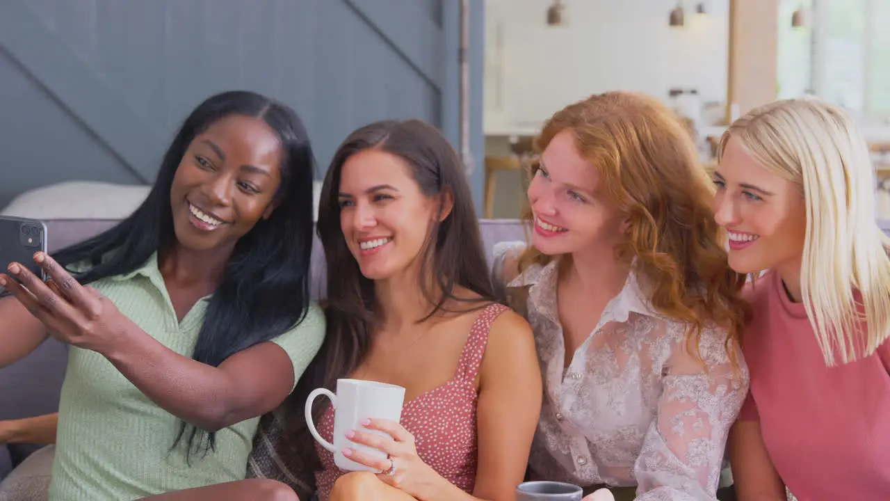 Multi-Cultural Group Of Female Friends Sitting On Sofa At Home Posing For Selfie Together